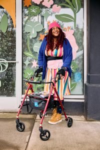 A non-binary Black woman stands in front of a shop with their sticker-covered rollator, smiling and looking off in the distance. Their colorful outfit includes a beanie hat, cardigan, waist trainer filled with ice packs, rainbow-striped dress, bike gloves layered over arthritis gloves, rainbow heart leggings, and sneakers.